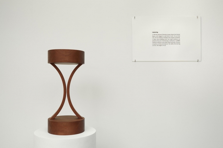 , IMAN ISSA&nbsp;Laboring (Study for 2012),&nbsp;2012&nbsp;Mahogany sculpture, text panel under glass, and white plinth Overall: 53 x 11 12 in. (134.62 x&nbsp;29.21 cm)&nbsp;Text panel: 11 x 17 in. (27.94 x 43.18 cm)&nbsp;Edition of 4&nbsp;Courtesy Rodeo Gallery